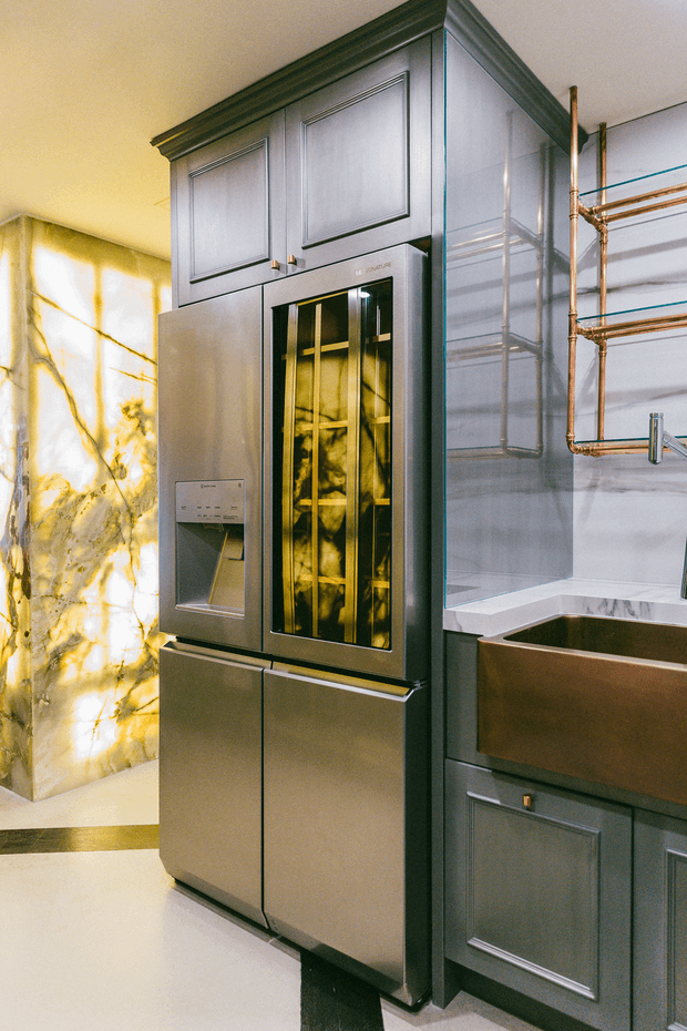 A luxurious, highly detailed kitchen interior featuring dark blue-grey paneled cabinetry, a tall integrated refrigerator or freezer with glass-front compartments, a deep copper-toned farmhouse sink, exposed copper plumbing, and dramatic backlit marble-effect wall panels. The design combines traditional panelled joinery with refined contemporary appliances and metallic accents.