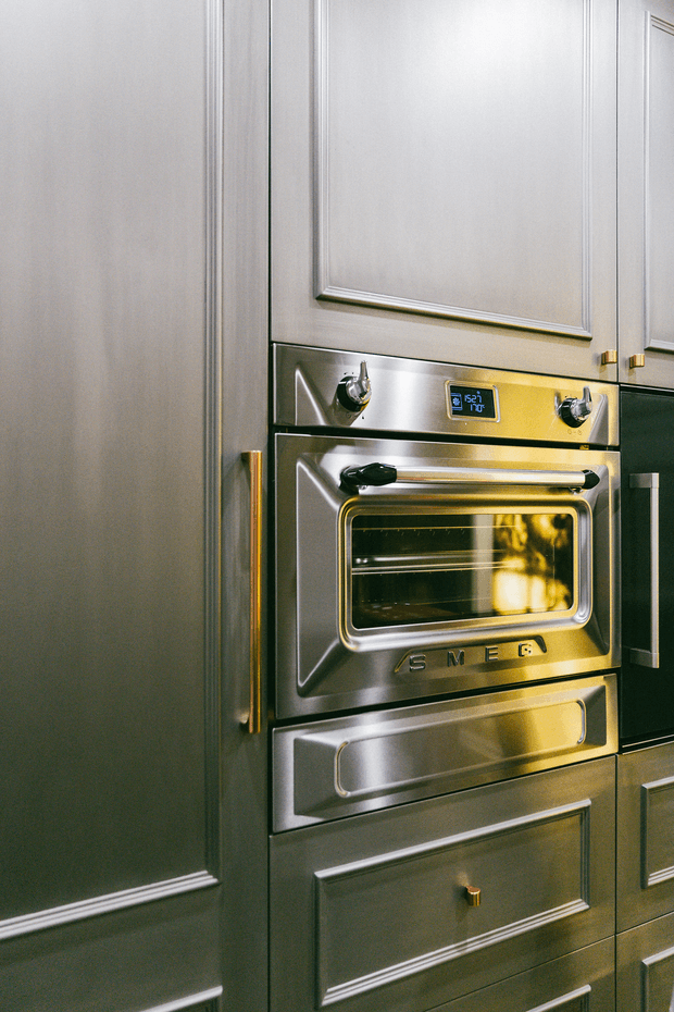 Close-up view of a built-in stainless-steel Smeg oven integrated into dark grey, framed cabinetry. The cabinetry features raised-panel doors and drawers with slim brass-gold handles, while the oven has a black glass window, metallic controls, and warm reflected light.