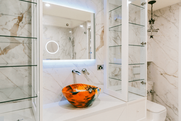 Luxurious contemporary bathroom featuring dramatic white marble-look wall finishes with warm veining, a floating white vanity with an artistic orange vessel basin, illuminated mirrored cabinetry, glass display shelving, and a partially enclosed shower area. Chrome fixtures, recessed lighting, and a concealed toilet contribute to the refined, high-contrast design.