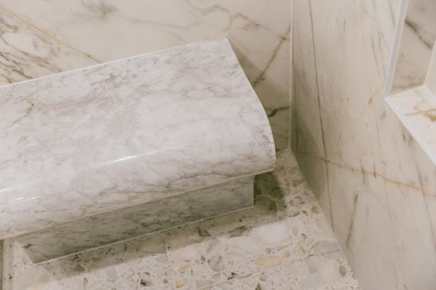 Close-up of a luxurious bathroom featuring a freestanding or built-in marble bathtub, marble wall cladding, and a pale terrazzo-style stone floor. The space has a cohesive light, veined stone finish with subtle warm undertones.
