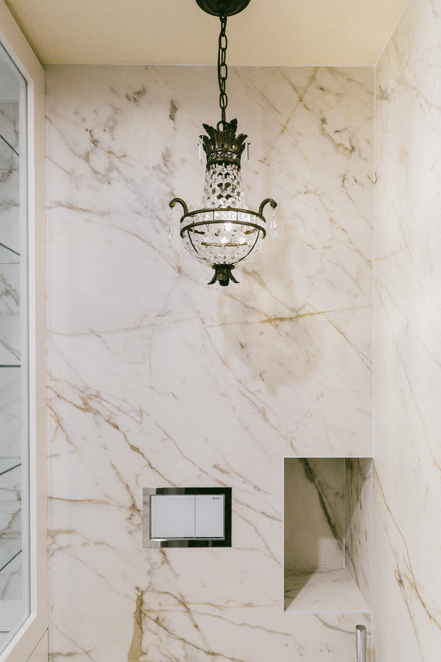 Luxurious bathroom interior with floor-to-ceiling white marble wall cladding featuring prominent grey and warm veining. An ornate hanging crystal chandelier provides a decorative focal point, while a minimalist dual-flush control and recessed wall niche add functional details. A frameless glass shower enclosure is partially visible at the left.