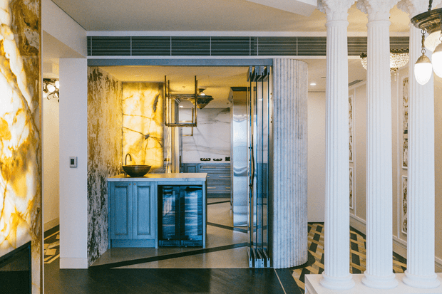 A refined open-plan kitchen and bar area framed by ornate fluted columns. The space features dramatic veined stone wall panels, a compact island with a dark bowl sink, blue-grey cabinetry, glass-front wine refrigeration, stainless-steel appliances, and a geometric dark-and-light tiled floor. Decorative classical detailing is combined with polished contemporary finishes and warm ambient lighting.