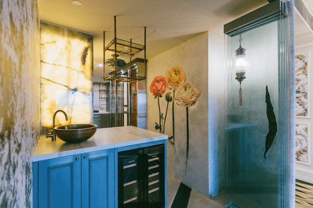 A richly decorated bathroom vanity area with bright blue built-in cabinetry, a pale stone benchtop, dark vessel basin and curved black tap. A mirrored shelving unit and patterned floral wall artwork add visual interest, while a frosted glass shower enclosure with a hanging pendant is visible to the right. The space uses warm lighting, expressive botanical motifs and a polished, eclectic decorative treatment.