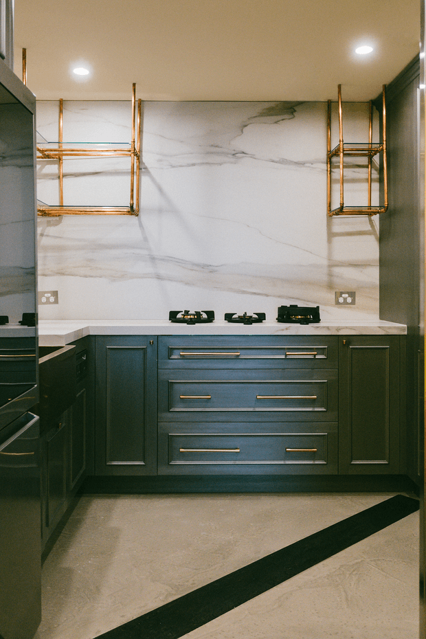 A compact, highly detailed kitchen featuring dark blue-green shaker-style cabinetry with brass bar pulls, a pale veined stone backsplash and benchtop, brass open shelving, and a built-in gas cooktop. The space has a warm, refined palette with recessed ceiling lighting and a pale floor crossed by a dark linear inlay or threshold.