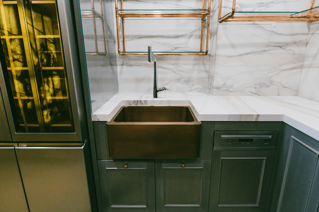 Close-up of a refined kitchen corner featuring a deep apron-front farmhouse sink, dark green-gray shaker cabinetry, a white veined stone benchtop and matching marble-look splashback. A tall dark-framed glass-front cabinet is visible at left, while exposed copper plumbing rails add a warm metallic accent above the counter.