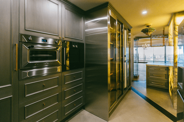 A narrow, highly finished kitchen or butler-style preparation area with dark built-in cabinetry, integrated stainless-steel oven and microwave, tall reflective pantry or storage cabinetry, and a central island visible toward the bright glazed end of the room. Gold-toned hardware, trim, and warm lighting create a dramatic, luxurious atmosphere.