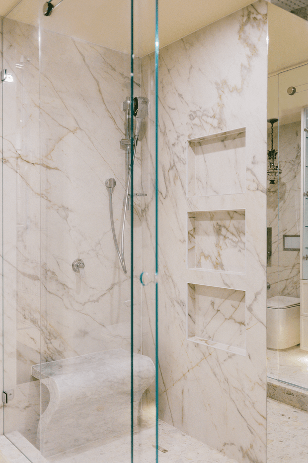 Luxurious walk-in shower with full-height white marble wall and floor surfaces featuring prominent grey and warm veining. Clear frameless glass panels and door enclose the shower, with a handheld and overhead shower fitting, a curved built-in marble bench, and three recessed marble-lined niches. A partial vanity area is visible through the glass at right.