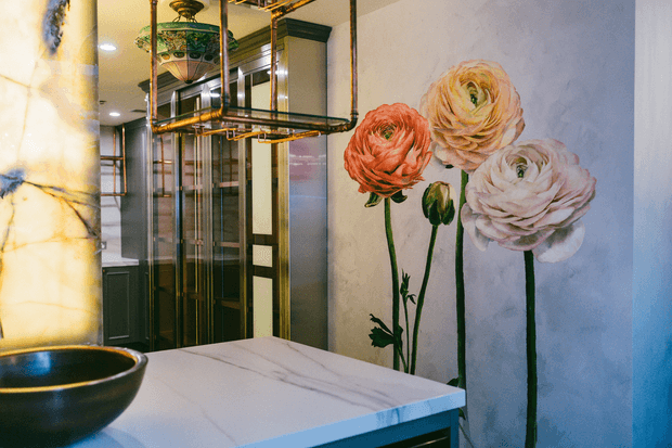 A richly detailed bathroom interior featuring a pale marble vanity benchtop with a dark vessel basin, a large floral wall mural depicting oversized ranunculus flowers, dark framed glass cabinetry or shower enclosure, and an ornate suspended metal-and-glass light fixture. The palette combines cool blue-grey walls with warm brass, black, cream, coral, and soft pink accents, creating a luxurious, art-deco-inspired atmosphere.