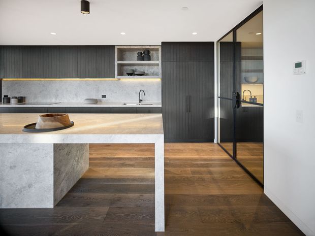 Contemporary kitchen with a large stone island, dark vertically ribbed cabinetry, warm timber flooring, integrated shelving, under-cabinet lighting, and black-framed glazed doors opening to an adjacent space.