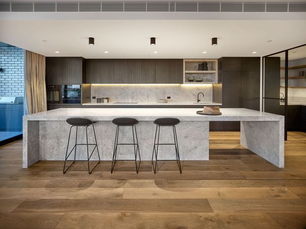 A sophisticated contemporary kitchen dominated by a large stone island with three black bar stools. Dark timber cabinetry spans the rear wall, paired with a pale stone backsplash and integrated appliances. Warm timber flooring, concealed under-cabinet lighting, open shelving, and black-framed glazing create a refined, understated interior.