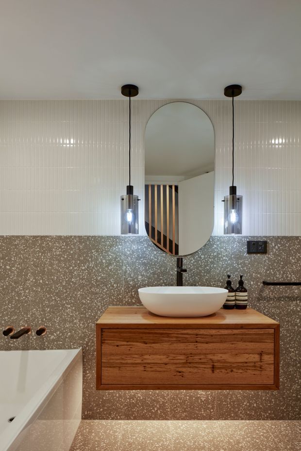 A contemporary bathroom vanity composition with a floating timber drawer cabinet, white vessel basin, arched oval mirror, and twin pendant lights. The walls combine vertically ribbed white tiles above and terrazzo-style speckled tiles below, with a partially visible freestanding bathtub and black tapware. The mirror reflects a timber staircase with dark and light vertical detailing.