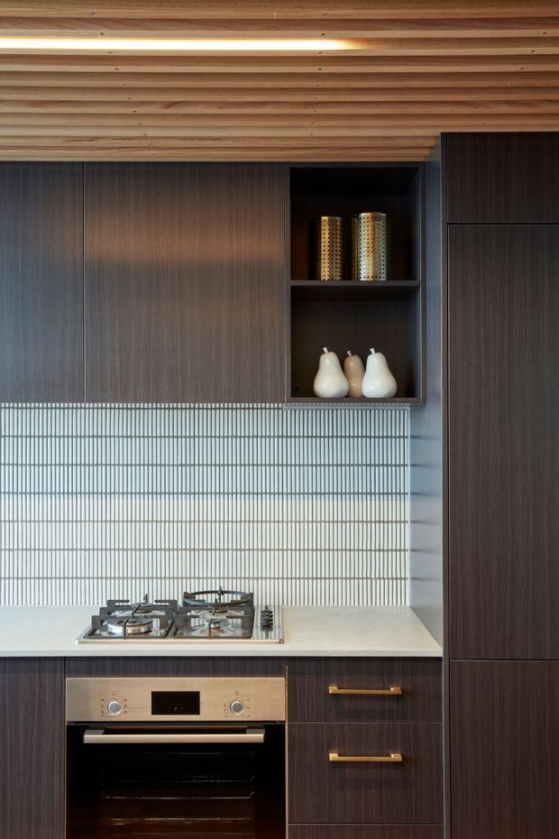 A compact, refined kitchen with dark wood-grain floor-to-ceiling cabinetry, integrated oven, four-burner gas cooktop, pale stone benchtop, and a vertically ribbed light tile splashback. An open display niche contains decorative ceramic vessels and metallic objects. The ceiling is lined with warm timber boards and includes a slim recessed light.