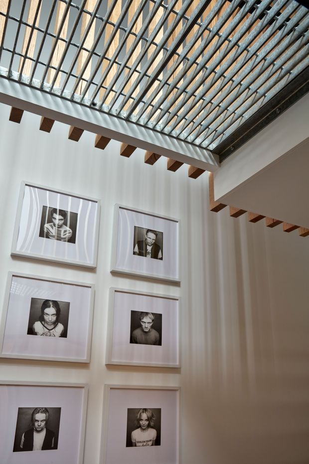 Interior view of a tall, light-filled gallery-like space with white walls, exposed dark timber beams, and a dramatic glazed roof structure incorporating repeated timber rafters and dark metal framing. Several framed black-and-white portrait photographs are arranged in a grid on the wall. Strong linear shadows from the roof glazing fall across the walls.