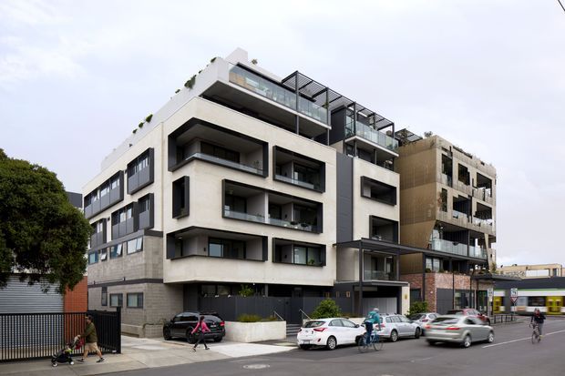 Street-level exterior view of a contemporary multi-storey apartment building with a stepped massing, pale rendered and concrete masonry facade, dark-framed windows, recessed balconies, glass balustrades, rooftop terraces, and planted roof edges. The building fronts an urban street with pedestrians, cyclists, parked cars, and neighbouring commercial frontage.