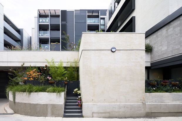 Contemporary multi-level residential development viewed from the street, featuring pale rendered or concrete walls, dark metal trims, recessed balconies, vertical screening, and landscaped raised planter beds with steps leading into the property.