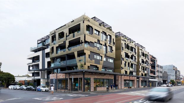 Street-level exterior view of a contemporary multi-storey mixed-use apartment building, with ground-floor commercial tenancies, upper-level balconies, extensive glazing, varied projecting volumes, and rooftop terraces. The building occupies an urban corner site alongside a marked city street.