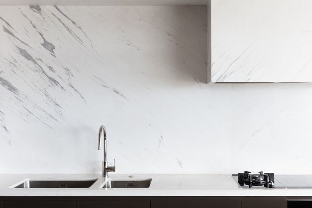 Minimal contemporary kitchen detail featuring a white marble-look stone splashback and benchtop with grey veining, an undermount double sink, chrome gooseneck tap, gas cooktop, and dark lower cabinetry.