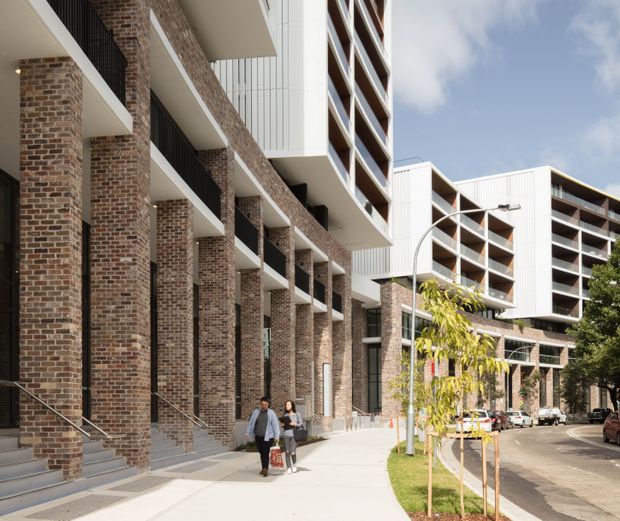 Street-level exterior view of a contemporary multi-storey apartment or mixed-use building complex. The facade combines repeated red-brown brick columns and piers with white projecting balconies, dark metal balustrades, and large recessed openings. A broad pedestrian sidewalk, young street trees, curved street lighting, and parked cars frame the urban setting.