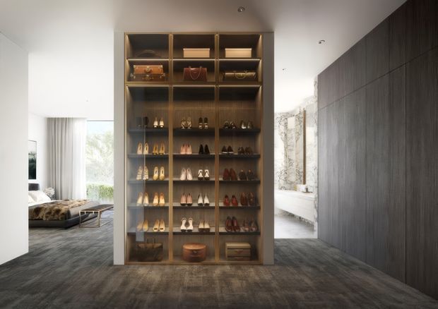 A refined walk-in wardrobe or dressing area centered on a tall, floor-to-ceiling open display cabinet with warm timber or bronze-toned framing and illuminated shelves holding shoes, handbags, and accessories. Dark vertical wall panelling lines the right side, while a bedroom is visible through the opening on the left and a bright bathroom with a freestanding tub appears beyond on the right. The space has dark textured carpet, white walls and ceiling, recessed spotlights, and a luxurious, gallery-like presentation.