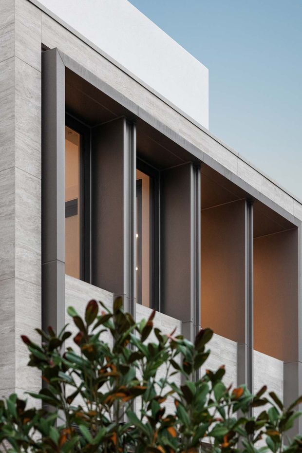Close-up exterior view of a contemporary multi-level facade with pale stone or concrete cladding, deep recessed balconies, dark vertical metal framing, and warm-lit glazing. Dense planting partially obscures the lower facade.