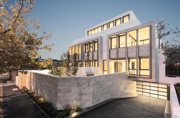 Exterior view of a multi-storey contemporary residence at dusk, featuring clean white rendered volumes, extensive floor-to-ceiling glazing, grey masonry wall panels, a projecting upper-level roof form, and an integrated basement garage accessed by a sloped driveway. Landscaping and boundary walls frame the facade.