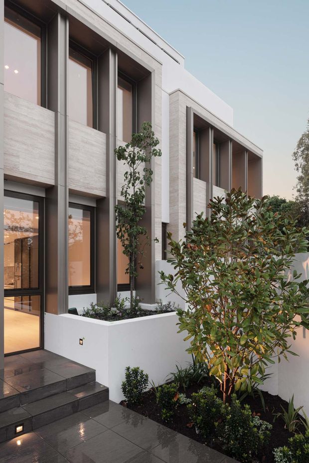 Contemporary multi-level residential facade viewed from the landscaped entry path. The building features pale textured masonry or stone cladding, deep recessed vertical window bays with dark metal frames, projecting geometric volumes, and a dark tiled staircase and terrace. Raised white planters and dense ornamental planting soften the crisp architectural lines.