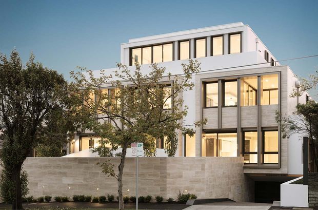 Contemporary multi-storey building facade with clean white rendered walls, extensive floor-to-ceiling glazing, dark window frames, and a projecting upper-level form. A light stone boundary wall and landscaped street frontage frame the entrance and lower-level garage opening.