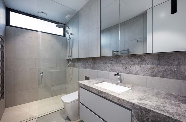 Contemporary bathroom with a walk-in tiled shower, wall-hung toilet, large mirrored cabinetry, and a floating vanity with a pale stone-look benchtop and integrated basin. The room has cool neutral finishes, dark trim, and natural light from a high horizontal window.