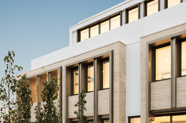 Close-up exterior view of a multi-storey contemporary building with a clean white rendered facade, pale stone-look vertical wall panels, and tall dark-framed windows. Warm interior lighting is visible through the glazing, with young trees in the foreground and a clear blue sky behind.