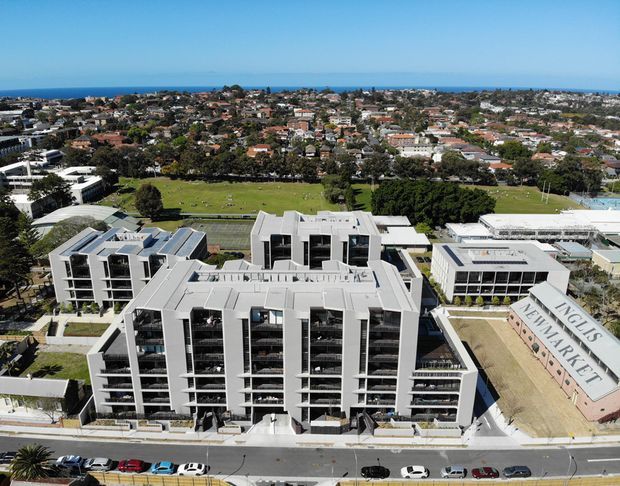 Aerial view of a contemporary multi-storey apartment or mixed-use development beside a large landscaped recreation field, with dense suburban housing and the ocean visible in the distance. The complex features flat roofs, light-coloured façades, extensive balconies, dark window glazing, and a prominent sloped wall reading “English Newmarket.”