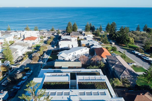 Aerial view of a low-rise coastal residential neighbourhood beside a deep blue ocean. The foreground shows a contemporary flat-roofed building with extensive rooftop glazing and planted roof areas, surrounded by detached homes, mature trees, streets, and parked vehicles. The image is dominated by the coastal setting and building rooftops.