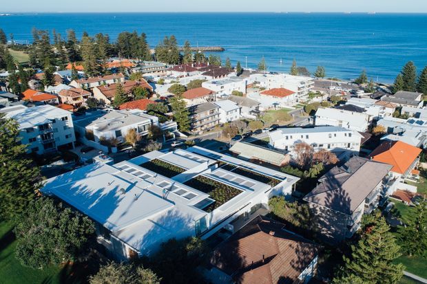Aerial view of a low-rise coastal residential neighbourhood beside a blue ocean, with a mix of detached homes and apartment buildings. The foreground includes a large contemporary flat-roof building with rooftop planted areas and skylights, while red-tiled and light-coloured pitched roofs are visible throughout the surrounding suburb. A tree-lined shoreline and small coastal pier appear in the background.
