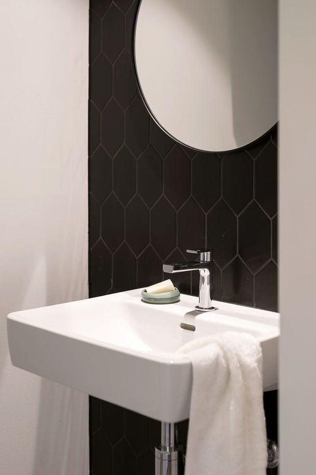 Close-up interior view of a compact bathroom vanity. A wall-mounted white rectangular basin with chrome tapware and exposed chrome plumbing sits against a black geometric elongated-hexagon tiled feature wall. A large round mirror is mounted above the basin, with a white towel hanging from the front edge. The scheme is monochromatic, clean, and contemporary with strong black-and-white contrast.
