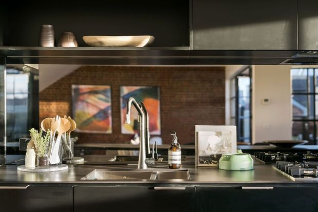 A contemporary kitchen viewed across a large island, featuring dark flat-panel cabinetry, a stainless-steel benchtop with integrated sinks, a tall curved mixer tap, gas cooktop, and open shelving with ceramic vessels. A mirrored splashback reflects exposed brickwork, artwork, and glazing from the adjoining living area, creating depth and a moody industrial character.