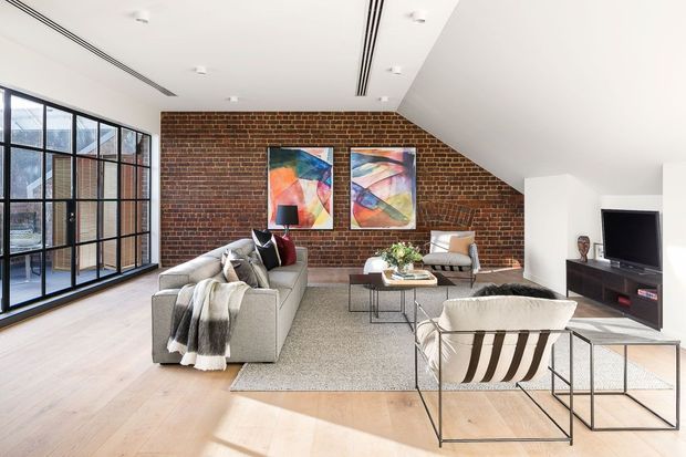 Bright contemporary living room with an exposed red-brick feature wall, sloping white ceiling, pale timber flooring, and expansive black-framed floor-to-ceiling glazing. The room is furnished with a neutral sectional sofa, lounge chairs, a low coffee table, rug, and media console, with colourful abstract artwork adding visual interest.