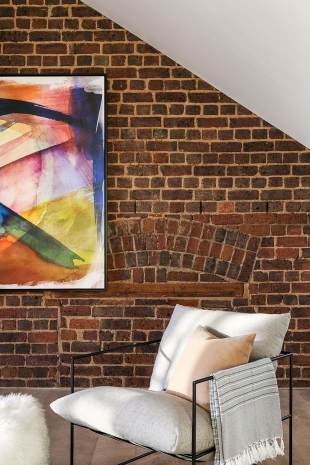 Close-up interior detail of a warm, industrial-style sitting area. An exposed red brick wall with a prominent arched brick feature forms the backdrop, contrasted by a sharply sloping white ceiling. A black metal-framed lounge chair with pale cushions and a draped striped throw sits on a timber floor, with a colourful abstract artwork mounted on the wall.