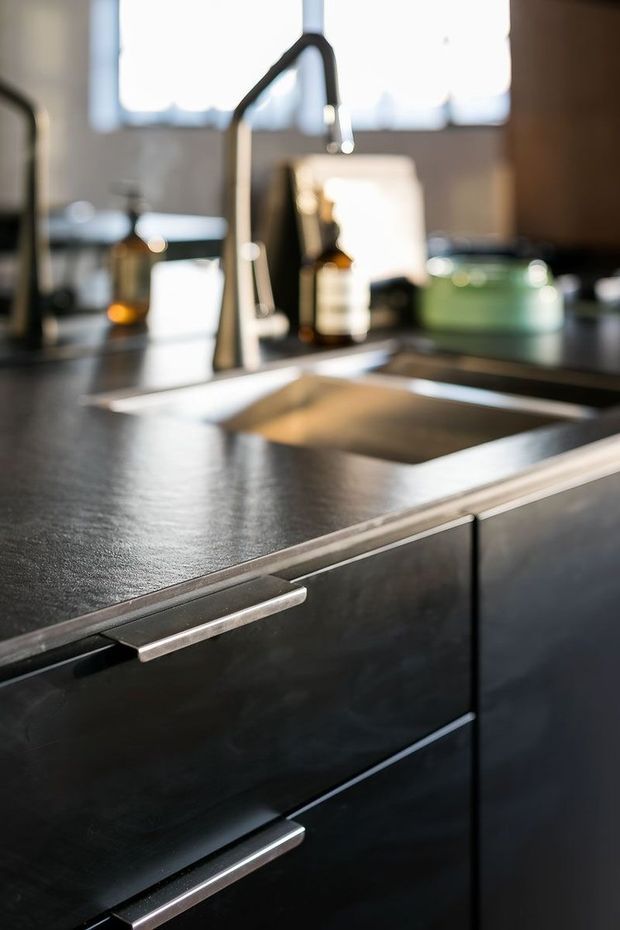 Close-up photograph of a contemporary kitchen centred on a deep stainless-steel sink and tall curved gooseneck tap. The sink is set into a dark, textured benchtop, with dark flat-panel cabinetry and long brushed-metal bar handles in the foreground. Soft daylight enters through a window behind the sink, while blurred kitchen accessories sit on the rear counter.