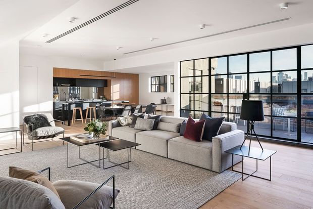 Bright, open-plan contemporary apartment interior combining a living room, dining area and kitchen. The room features a large light-grey sectional sofa, black metal-framed coffee tables, neutral textured rug, timber flooring, a black-framed glazed wall with city views, and a kitchen with warm timber cabinetry and a dark benchtop. Recessed lighting and linear ceiling vents contribute to the clean, modern feel.