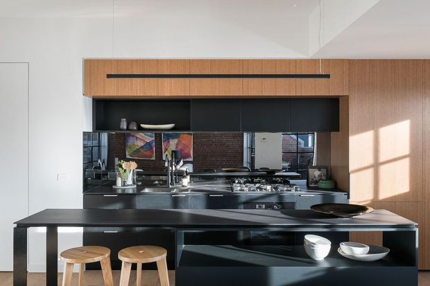 A contemporary kitchen with a large dark island bench and integrated seating, black cabinetry and benchtops, a warm timber overhead cabinet, and a dark brick backsplash. The space has white walls, strong natural light, and a restrained, high-contrast palette.