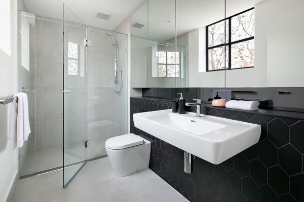 A bright contemporary bathroom with a frameless glass walk-in shower, wall-mounted white toilet, and a large white basin set into a dark charcoal vanity. The walls and floor are finished in pale grey, while the vanity wall features dark geometric hexagonal tiles and a slim black shelf. Large black-framed windows, mirrored surfaces, and minimal black fixtures complete the clean, modern scheme.