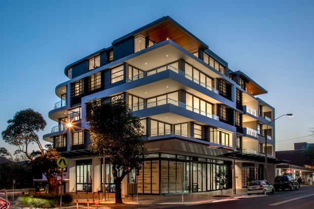Twilight exterior view of a contemporary multi-storey mixed-use apartment building on a street corner. The building features stacked balconies, extensive floor-to-ceiling glazing, dark horizontal cladding and screens, projecting roof forms, warm interior lighting, and a glazed ground-floor commercial frontage. Street trees, signage, parked vehicles, and surrounding urban context are visible.