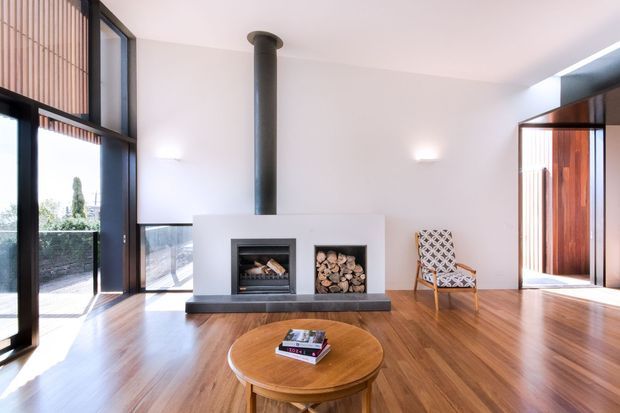 Bright, open-plan living room with polished timber flooring, expansive dark-framed glazing, a central double-sided or built-in fireplace with cylindrical flue, and a built-in log storage niche. A patterned lounge chair and round timber coffee table furnish the space. Vertical timber screening and a timber-clad entry door add architectural warmth, while white walls and high glazing create an airy contemporary interior.