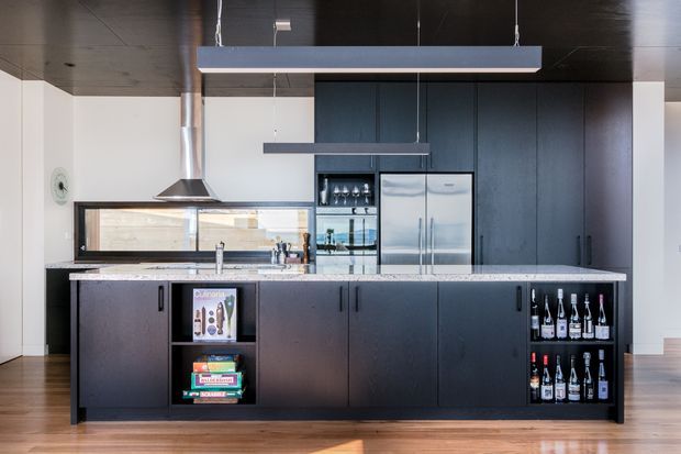 Contemporary kitchen featuring a large dark island with integrated open bottle shelving, pale stone benchtops, extensive dark cabinetry, stainless steel appliances, a black ceiling, timber flooring, and suspended rectangular pendant lights. A stainless steel rangehood and reflective splashback/window area are visible behind the island.