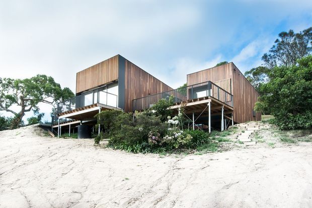 A modern two-volume timber-clad house elevated on slender supports above a sandy, sloping site. Large glazed openings, projecting balconies and decks, and exposed structural posts create a lightweight connection to the surrounding coastal-looking landscape. Native vegetation surrounds the building, with a stepped path leading up to the right-hand volume.