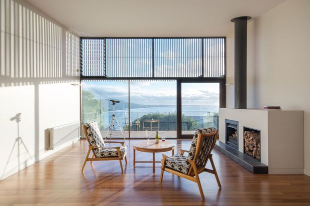 Bright, airy contemporary living room with warm timber flooring, a minimalist white fireplace and tall black flue, and expansive glazing opening to a scenic coastal or waterfront view. Vertical external or internal blinds cast strong linear shadows across the walls, while two patterned timber lounge chairs and a round coffee table create a relaxed seating area.