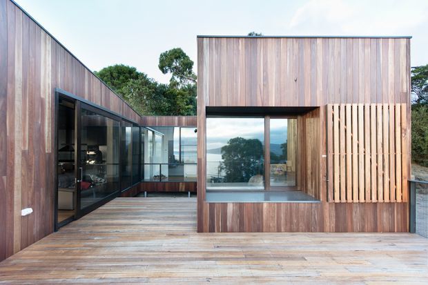 Exterior view of a modern timber-clad residence arranged around a broad timber deck and courtyard-like outdoor space. The building has clean rectilinear volumes, extensive floor-to-ceiling glazing, a projecting upper-level box, and vertical timber screening. Water and distant landscape are visible beyond the glazing.
