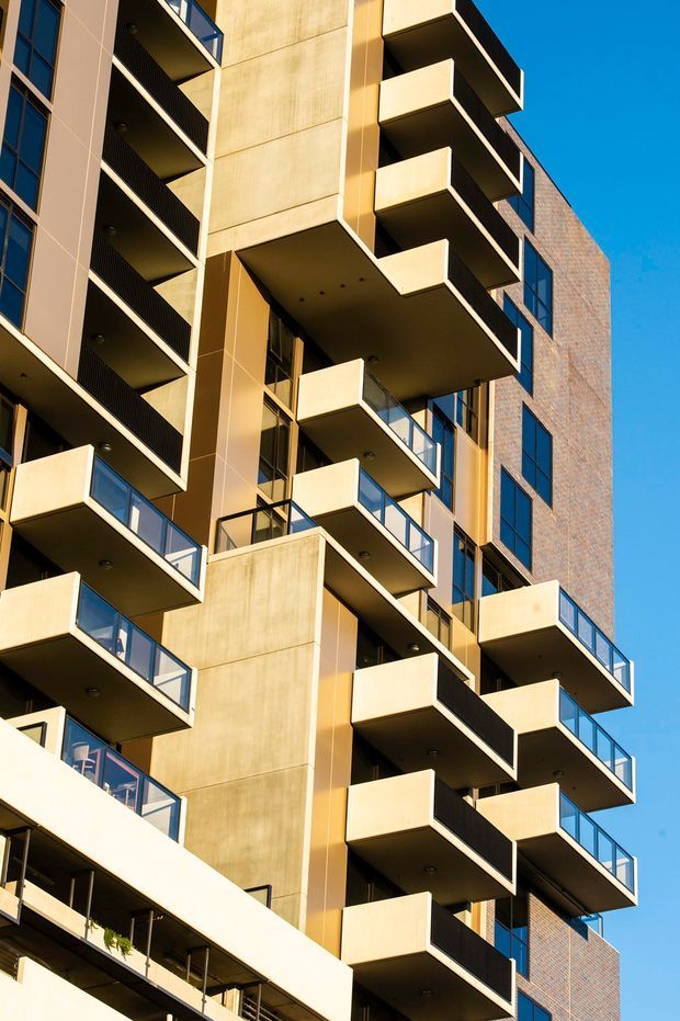Close-up upward-looking view of a contemporary multi-storey apartment building facade, dominated by projecting balconies with solid cream-coloured parapets, dark undersides, and blue-tinted glass balustrades. The facade combines smooth concrete or rendered panels with warm brickwork and extensive dark-framed glazing against a clear blue sky.