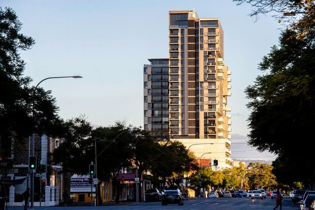 Street-level exterior view of a tall contemporary residential apartment building rising above a tree-lined urban street. The building has a multi-storey form with stacked balconies, extensive floor-to-ceiling glazing, dark vertical facade elements, and warm light-coloured projecting frames. Shops, traffic signals, parked vehicles, and mature street trees occupy the foreground.