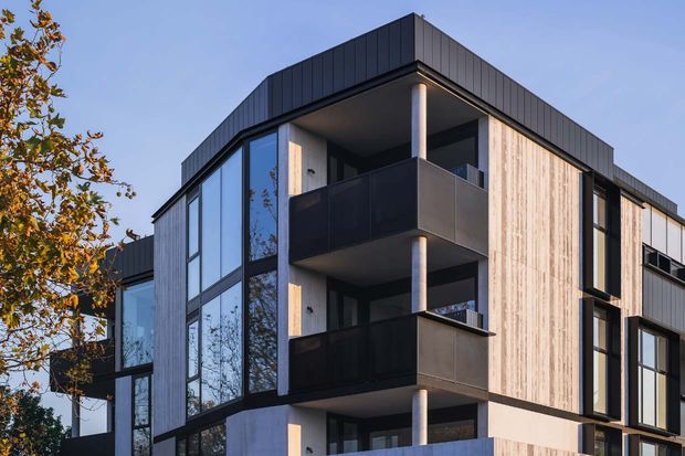 Exterior view of a contemporary multi-storey residential building with a strong geometric form, dark standing-seam cladding, pale vertical timber-look wall panels, extensive black-framed glazing, and stacked recessed balconies with dark balustrades.