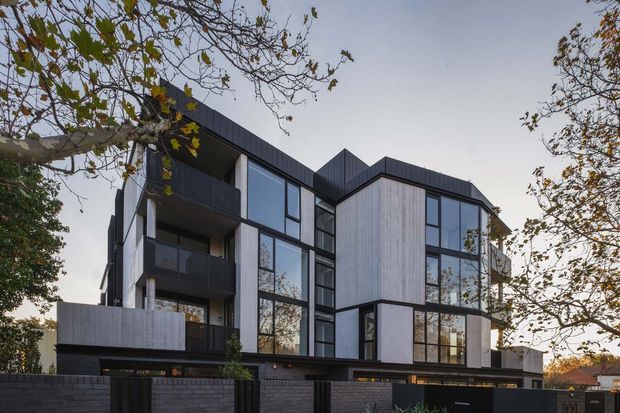 Contemporary multi-storey residential building viewed from the street, with angular roof forms, dark metal cladding and framing, large glazed windows, projecting balconies, and light grey vertical wall panels. A dark masonry boundary wall and mature deciduous trees frame the facade.